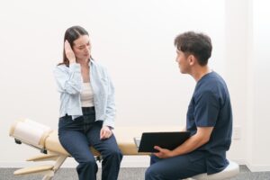 頭に手を当ててつらさを話す女性の話を、施術者がバインダーを持って聞いている様子