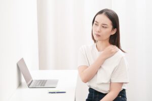 机の前に座り、肩に手を当ててつらそうにしている女性