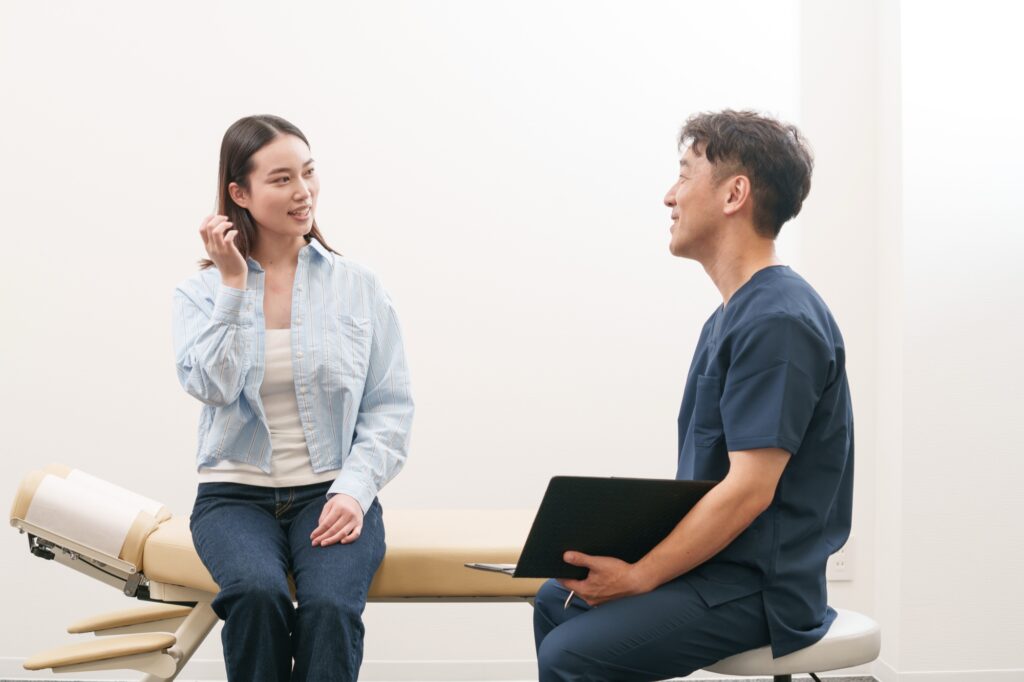 施術ベッドに座る女性が笑顔で話し、施術者がバインダーを持って聞いている様子
