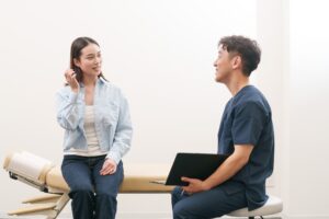 施術ベッドに座る女性が笑顔で話し、施術者がバインダーを持って聞いている様子